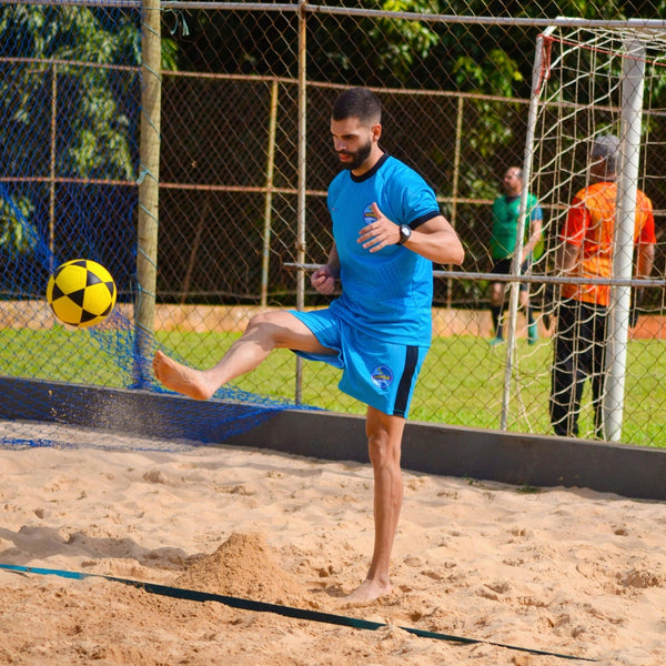 Athletic man in blue jersey kicking a yellow ball on a sandy beach soccer court