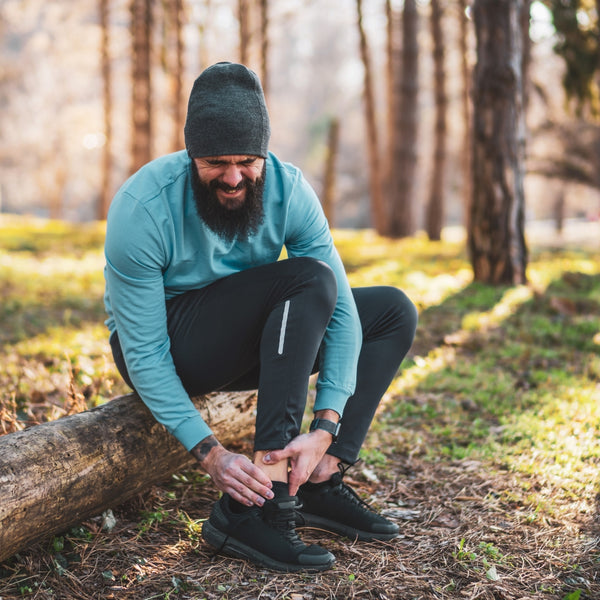 Bearded man in teal shirt tying black trail running shoes on a forest log