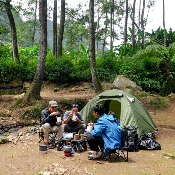 Three people camping in a forest, sitting around a green tent with cooking equipment.